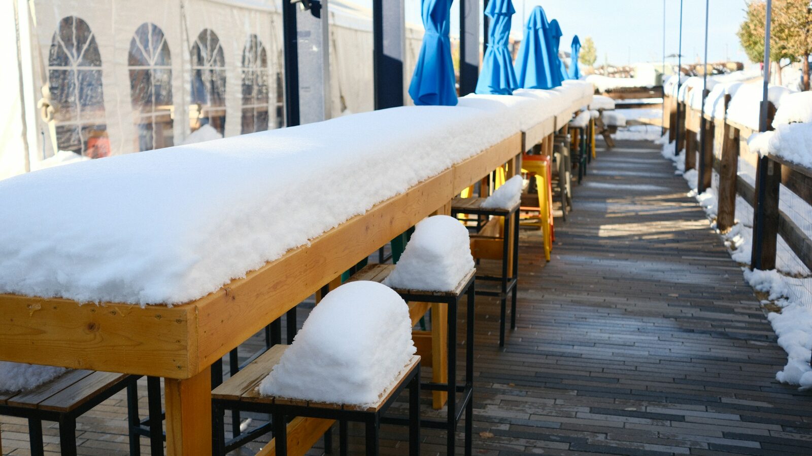 Winter sunshine on a long outdoor bar and stools covered in fresh snow, with blue umbrellas folded behind