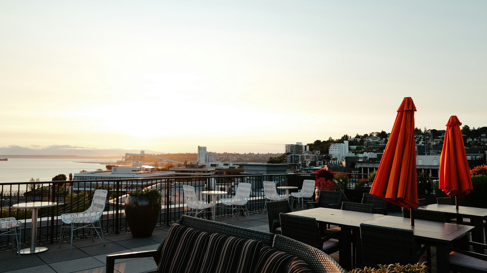 A rooftop terrace with red umbrellas and outdoor tables overlooking a city and bay as the sun sets