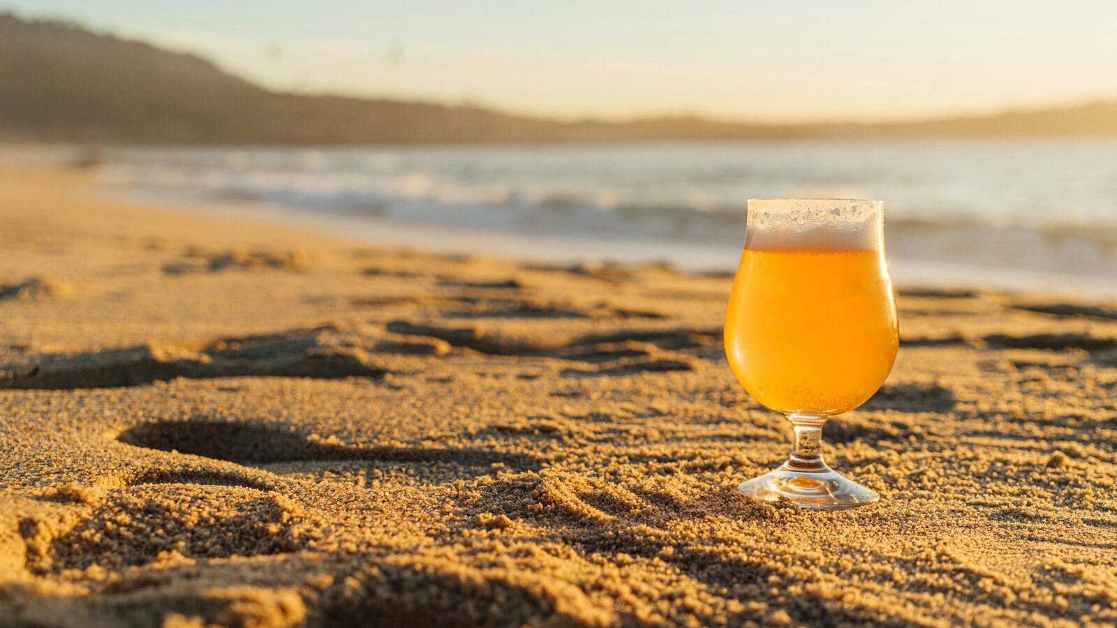 A glass of beer standing in the sand on a beach in warm, late afternoon sunshine