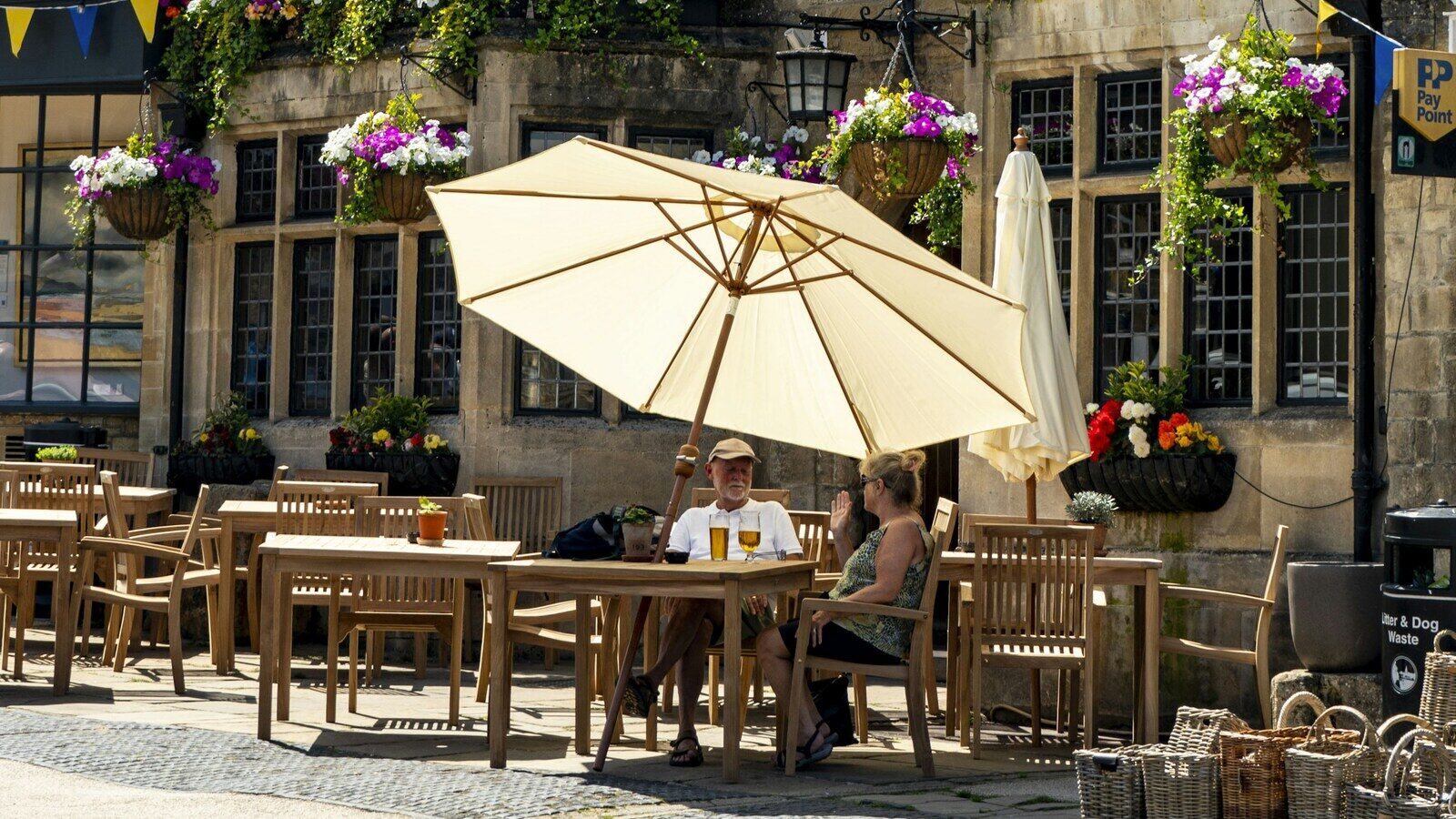 A couple sitting under a large umbrella at wooden tables outside a stone pub decorated with hanging flower baskets