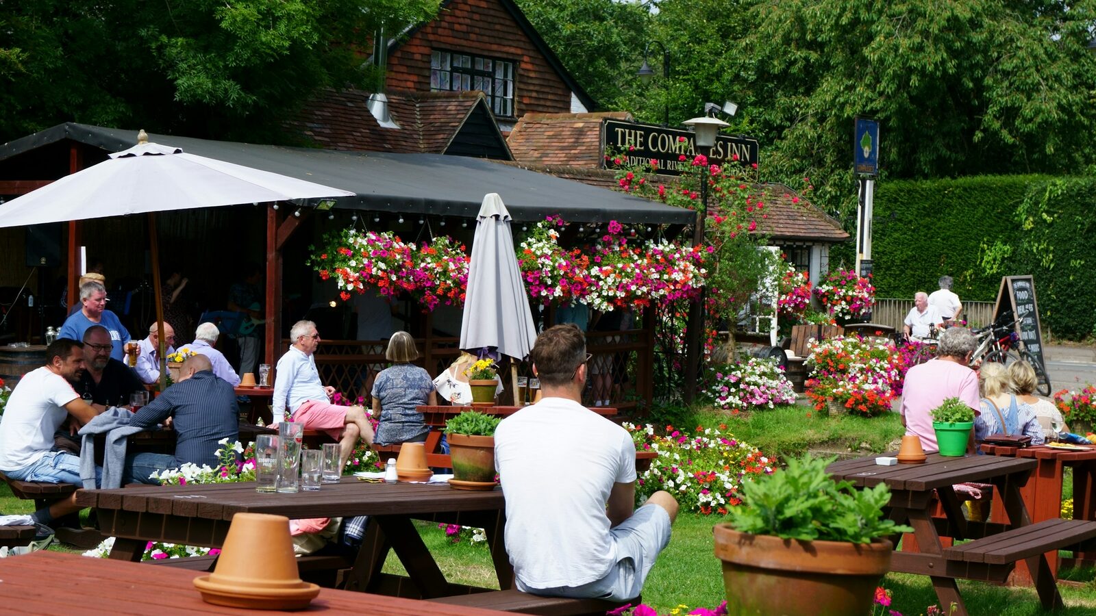People sitting at wooden picnic tables in a sunny village pub garden full of flowers