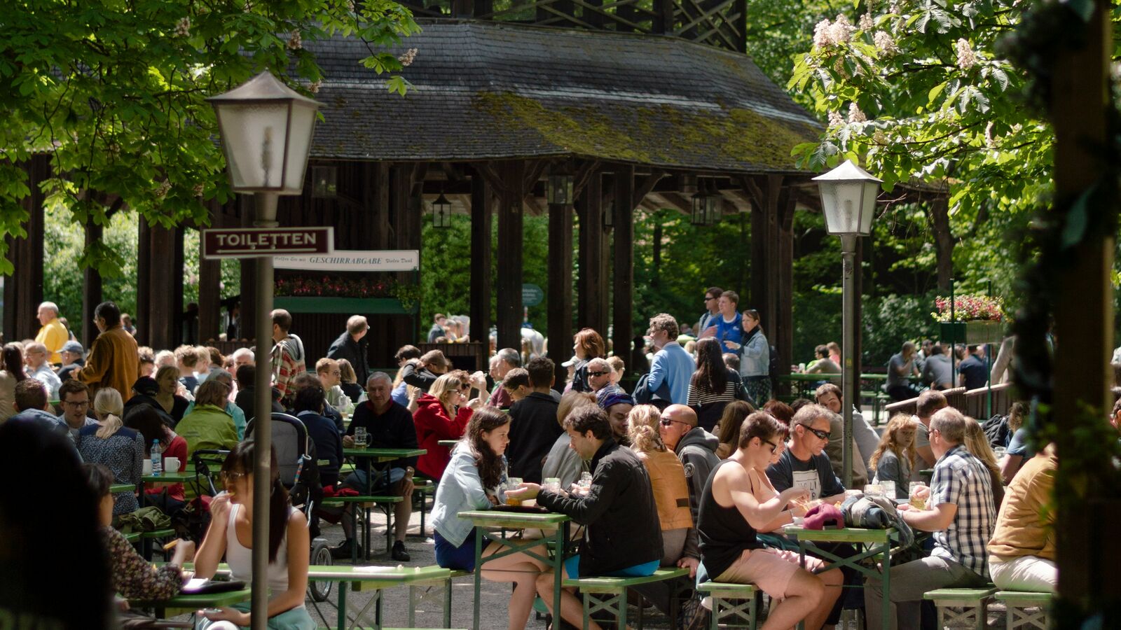 Crowds of people at wooden tables under leafy green trees in a sunny Munich beer garden
